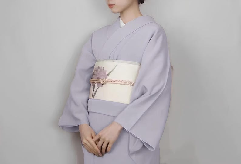 May include: A woman wearing a light purple kimono with a white obi featuring a floral design. The obi is tied with a decorative knot.