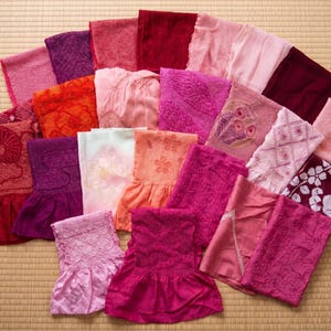 May include: A collection of colorful, folded textiles in shades of pink, red, and purple. Some pieces feature intricate patterns and floral designs. The items are arranged on a light brown, woven surface.