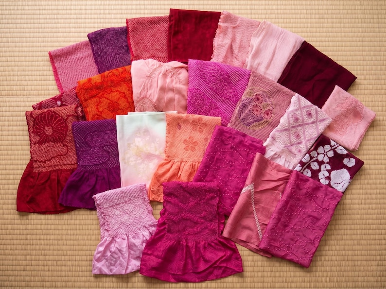 May include: A collection of colorful, folded textiles in shades of pink, red, and purple. Some pieces feature intricate patterns and floral designs. The items are arranged on a light brown, woven surface.