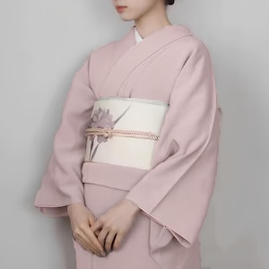 May include: A pale pink kimono with a white obi featuring a floral design. The kimono has long sleeves and a traditional Japanese style.