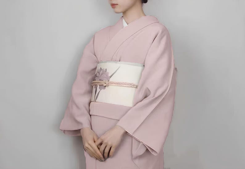 May include: A pale pink kimono with a white obi featuring a floral design. The kimono has long sleeves and a traditional Japanese style.