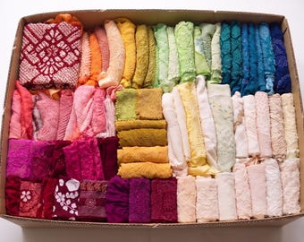 May include: A cardboard box filled with a variety of colorful fabric scraps. The fabrics are arranged in rows and columns, showcasing a spectrum of colors from red to blue. The textiles appear to be of different textures and patterns.