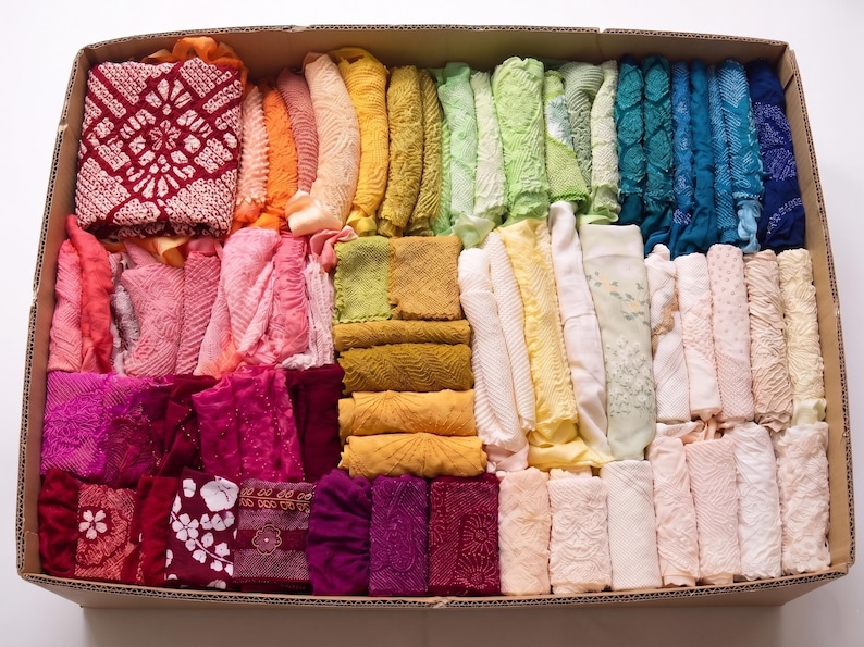 May include: A cardboard box filled with a variety of colorful fabric scraps. The fabrics are arranged in rows and columns, showcasing a spectrum of colors from red to blue. The textiles appear to be of different textures and patterns.