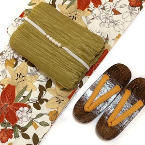 K&ouml;nnte beinhalten: Ein olivgr&uuml;nes, strukturiertes Stoffst&uuml;ck mit wei&szlig;em Band und Perlenakzenten, das auf einem Stoff mit Blumenmuster mit roten und gelben Lilien liegt. Ein Paar h&ouml;lzerne Geta-Sandalen mit orangefarbenen Riemen ist ebenfalls vorhanden.