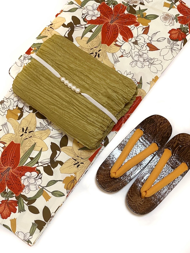 K&ouml;nnte beinhalten: Ein olivgr&uuml;nes, strukturiertes Stoffst&uuml;ck mit wei&szlig;em Band und Perlenakzenten, das auf einem Stoff mit Blumenmuster mit roten und gelben Lilien liegt. Ein Paar h&ouml;lzerne Geta-Sandalen mit orangefarbenen Riemen ist ebenfalls vorhanden.