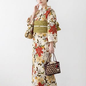 K&ouml;nnte beinhalten: Cremefarbener Kimono mit einem Blumenmuster aus roten und gelben Bl&uuml;ten. Der Kimono ist mit einem gr&uuml;nen Obi-G&uuml;rtel gebunden. Die Person h&auml;lt eine braune gewebte Handtasche und tr&auml;gt Holzsandalen.