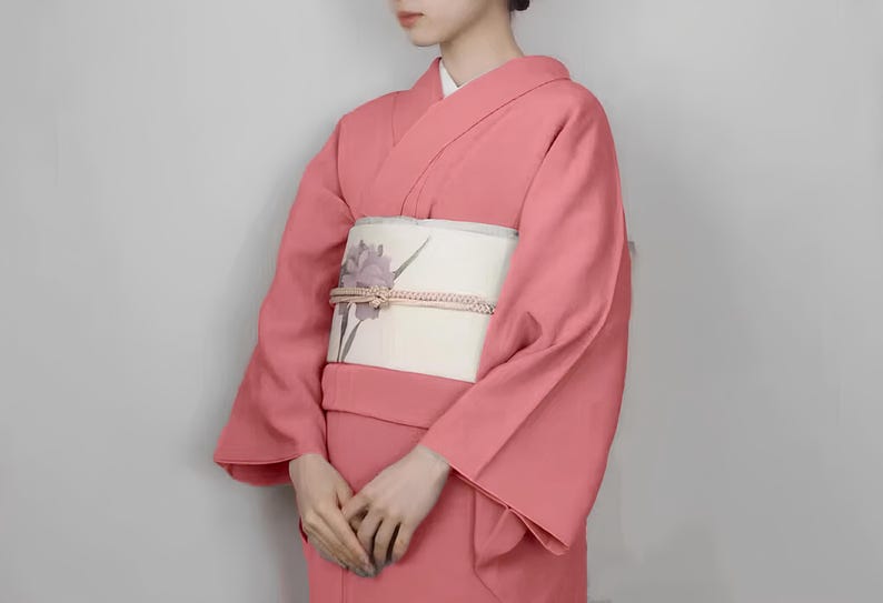 May include: A pink kimono with a white obi featuring a floral design. The kimono is made of a soft, flowing fabric and has a traditional Japanese style.
