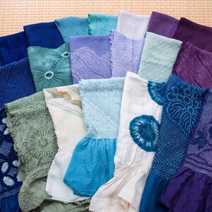 May include: A collection of colorful, patterned textiles in shades of blue, green, purple, and white. The fabrics feature various textures and designs, including tie-dye and embroidered patterns. The textiles are arranged on a light brown surface.
