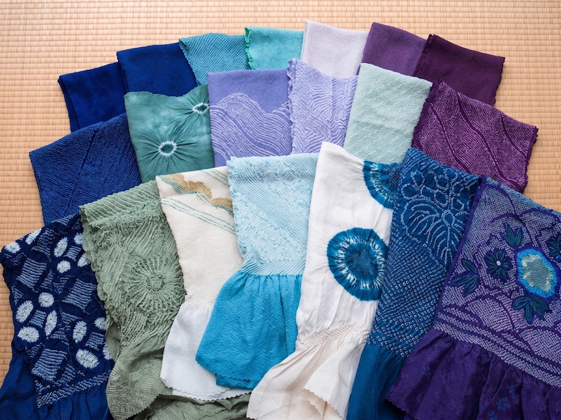 May include: A collection of colorful, patterned textiles in shades of blue, green, purple, and white. The fabrics feature various textures and designs, including tie-dye and embroidered patterns. The textiles are arranged on a light brown surface.
