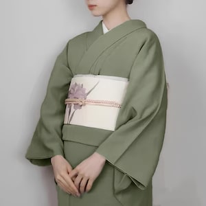 May include: A woman wearing a sage green kimono with a white obi featuring a floral design. The obi is tied with a decorative knot.