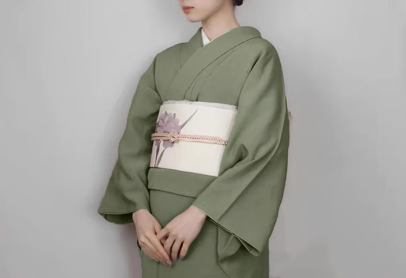 May include: A woman wearing a sage green kimono with a white obi featuring a floral design. The obi is tied with a decorative knot.