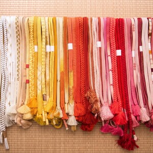 May include: A collection of colorful Japanese obi belts, displayed in a row. The belts vary in color, including white, yellow, orange, red, and pink. Some belts have tassels. The background is a woven mat.