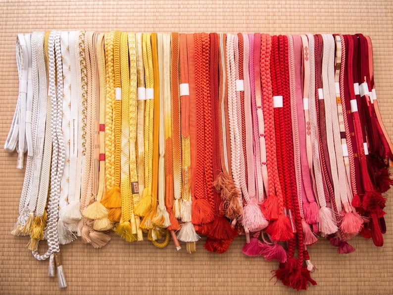 May include: A collection of colorful Japanese obi belts, displayed in a row. The belts vary in color, including white, yellow, orange, red, and pink. Some belts have tassels. The background is a woven mat.