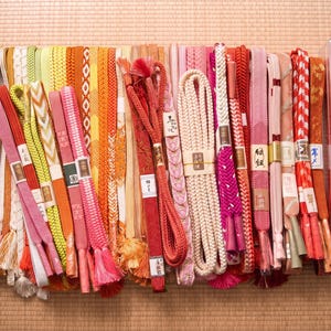 May include: A collection of colorful Japanese obi belts in various shades of red, pink, orange, yellow, and white. The belts are made of woven fabric and feature intricate patterns and tassels. Some are bundled with paper labels. The collection is displayed on a woven mat.