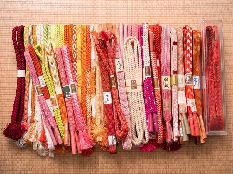 May include: A collection of colorful Japanese obi belts in various shades of red, pink, orange, yellow, and white. The belts are made of woven fabric and feature intricate patterns and tassels. Some are bundled with paper labels. The collection is displayed on a woven mat.