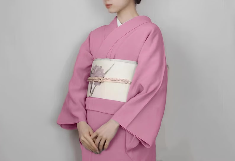 May include: A pink kimono with a white obi featuring a floral design. The kimono has long sleeves and a traditional style.