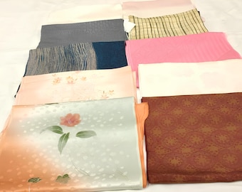 May include: A collection of folded fabric pieces in various colors and patterns. The fabrics include shades of pink, blue, green, and brown. Some pieces feature floral designs, while others have textured or geometric patterns. The fabrics are arranged on a white surface.