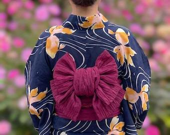 May include: A dark blue yukata featuring a pattern of orange and white goldfish and thin white wavy lines. The garment is tied at the waist with a wide, textured, deep burgundy obi sash that is folded into a large, decorative bow at the back.
