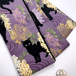 Puede incluir: Tela morada con un patrón de gato negro y flor de glicinia. La tela tiene un borde negro y está doblada por la mitad.