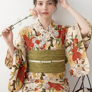 K&ouml;nnte beinhalten: Eine Frau tr&auml;gt einen cremefarbenen Blumenkimono mit roten und gelben Akzenten, kombiniert mit einem gr&uuml;nen Obi-G&uuml;rtel. Der Kimono hat ein gro&szlig;es Blumenmuster. Sie h&auml;lt eine Blume und einen geflochtenen Korb.