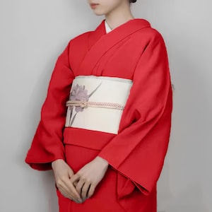 May include: A woman wearing a red kimono with a white obi featuring a floral design. The kimono has long sleeves and a traditional style.
