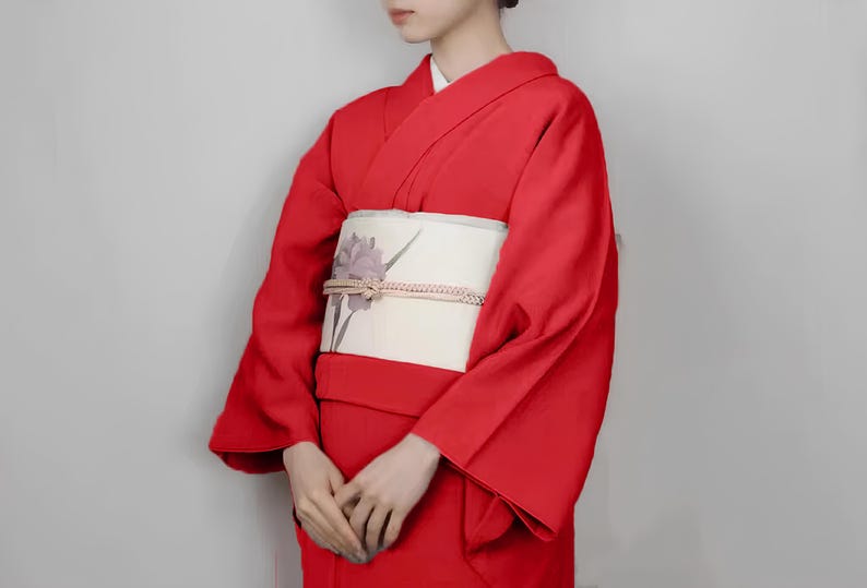 May include: A woman wearing a red kimono with a white obi featuring a floral design. The kimono has long sleeves and a traditional style.