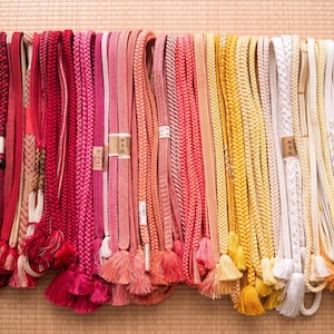 May include: A collection of colorful Japanese obi cords, displayed in a row. The cords vary in shades of red, pink, orange, yellow, and white. Many have tassels at the bottom. The cords are made of braided and woven materials.