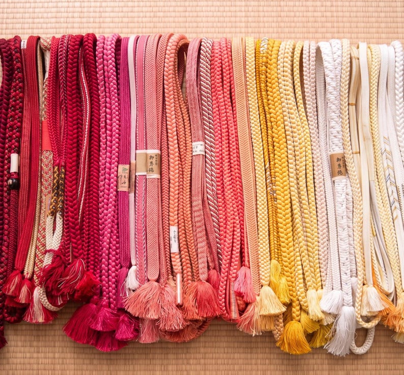 May include: A collection of colorful Japanese obi cords, displayed in a row. The cords vary in shades of red, pink, orange, yellow, and white. Many have tassels at the bottom. The cords are made of braided and woven materials.