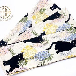 Puede incluir: Una tela blanca con un patrón repetido de gatos negros, flores azules, rosas y amarillas. La tela tiene un ligero brillo dorado.