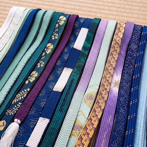 May include: A collection of colorful, patterned Japanese obi sashes. The sashes vary in shades of blue, green, purple, and gold, with intricate designs and textures. Some have tassels. The sashes are arranged on a woven mat.