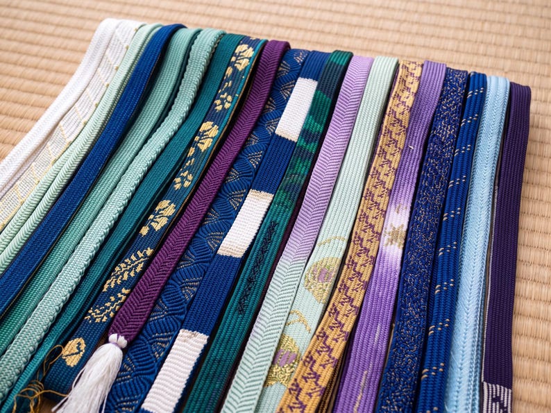 May include: A collection of colorful, patterned Japanese obi sashes. The sashes vary in shades of blue, green, purple, and gold, with intricate designs and textures. Some have tassels. The sashes are arranged on a woven mat.