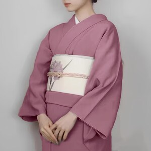 May include: A woman wearing a pink kimono with a white obi featuring a floral design. The kimono has long sleeves and a traditional style.