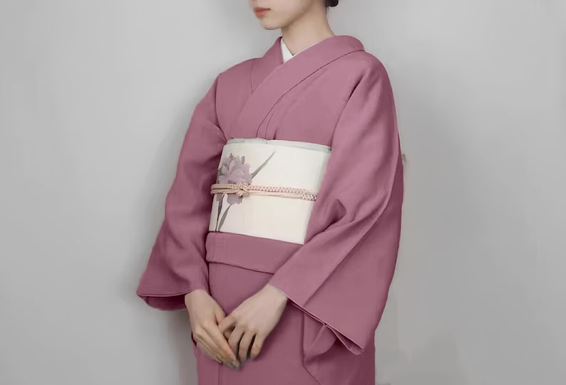 May include: A woman wearing a pink kimono with a white obi featuring a floral design. The kimono has long sleeves and a traditional style.