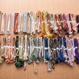 May include: A collection of colorful Japanese obi jime cords, displayed on a woven mat. The cords vary in color, including shades of green, pink, blue, and purple, and feature decorative tassels and intricate braiding. Some are packaged in boxes.