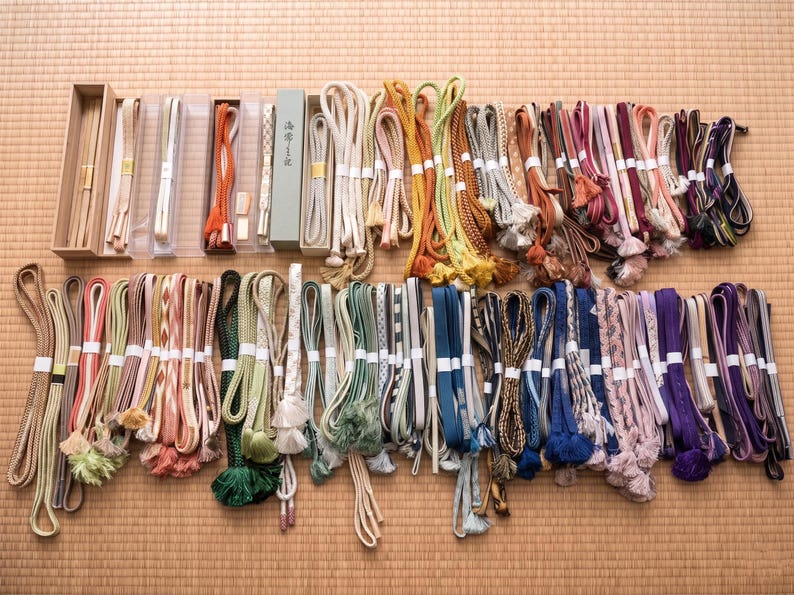May include: A collection of colorful Japanese obi jime cords, displayed on a woven mat. The cords vary in color, including shades of green, pink, blue, and purple, and feature decorative tassels and intricate braiding. Some are packaged in boxes.