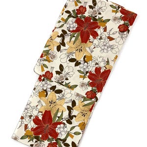 K&ouml;nnte beinhalten: Cremefarbener Stoff mit floralem Muster, das gro&szlig;e rote und gelbe Lilien, wei&szlig;e Bl&uuml;ten und gr&uuml;ne Bl&auml;tter zeigt. Der Stoff ist gefaltet und zeigt das detaillierte Muster. Das Design hat eine Vintage-&Auml;sthetik.