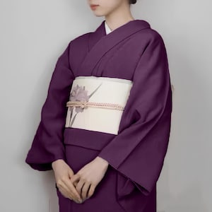May include: A woman wearing a purple kimono with a white obi featuring a floral design. The kimono has long sleeves and a traditional style.