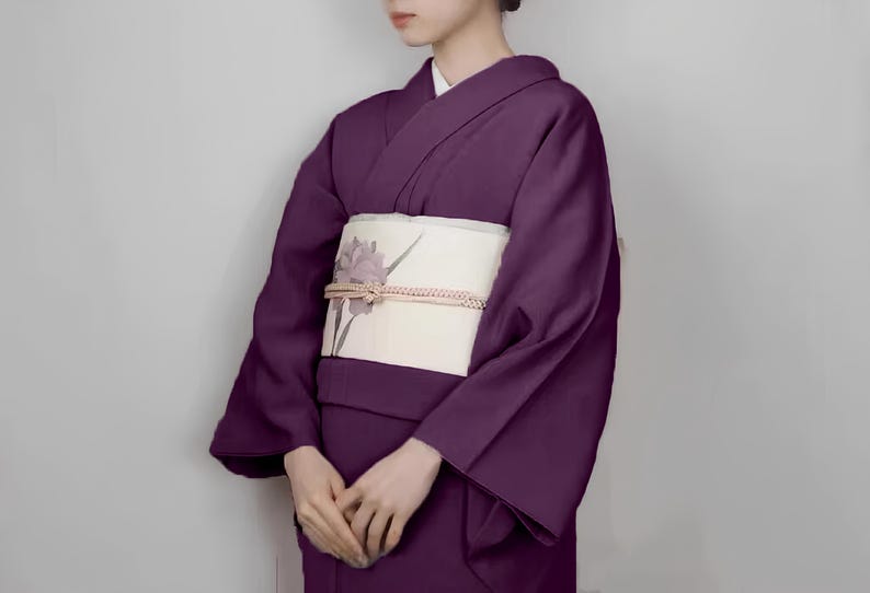 May include: A woman wearing a purple kimono with a white obi featuring a floral design. The kimono has long sleeves and a traditional style.