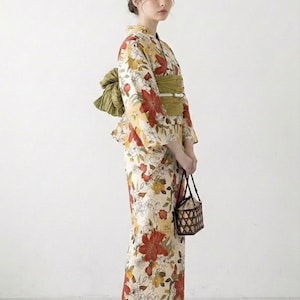 K&ouml;nnte beinhalten: Eine Frau in einem traditionellen japanischen Yukata. Das cremefarbene Kleidungsst&uuml;ck ist mit gro&szlig;en roten und gelben Blumenmustern verziert. Ein gr&uuml;ner Obi-G&uuml;rtel und eine passende Schleife betonen die Taille. Sie tr&auml;gt einen kleinen geflochtenen Korb und Holzsandalen.