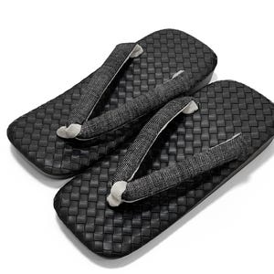 May include: A pair of black geta sandals. The sandals have a woven texture on the soles and feature fabric straps in black and white. The thong straps are attached to the sole with a white fabric.
