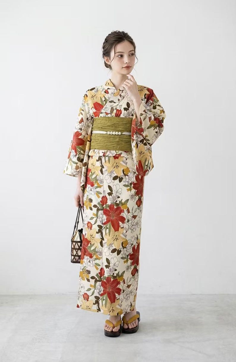 K&ouml;nnte beinhalten: Cremefarbener Kimono mit einem Blumenmuster aus roten und gelben Bl&uuml;ten. Der Kimono wird mit einem gr&uuml;nen Obi-G&uuml;rtel in der Taille zusammengehalten. Die Person tr&auml;gt Holzsandalen und eine kleine schwarze Tasche.
