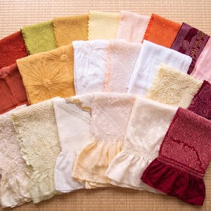 May include: A collection of colorful, folded textiles, including various shades of red, orange, yellow, green, and white. Some textiles feature embroidered or textured designs, while others have ruffled edges. The items are arranged on a light brown surface.