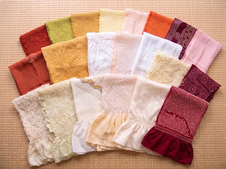 May include: A collection of colorful, folded textiles, including various shades of red, orange, yellow, green, and white. Some textiles feature embroidered or textured designs, while others have ruffled edges. The items are arranged on a light brown surface.
