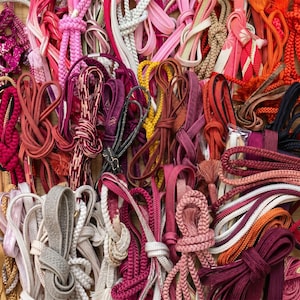 May include: A collection of colorful cords and ribbons in various shades of pink, red, orange, and white. The cords are knotted, braided, and looped, creating a textured and visually rich display. Some have decorative tassels.