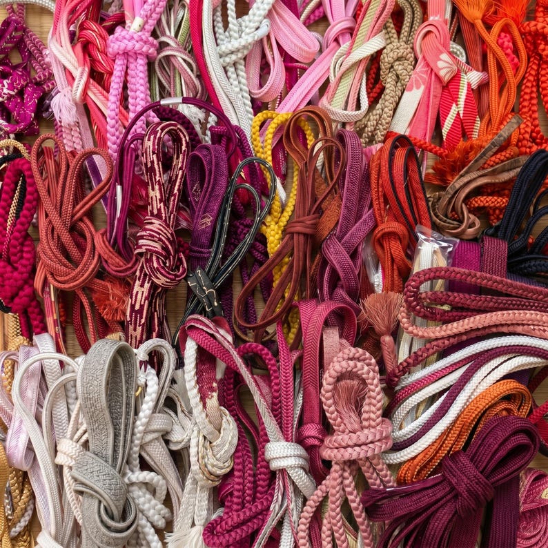 May include: A collection of colorful cords and ribbons in various shades of pink, red, orange, and white. The cords are knotted, braided, and looped, creating a textured and visually rich display. Some have decorative tassels.