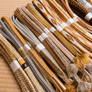 May include: A collection of intricately woven decorative cords in various shades of gold, silver, and beige. Some cords have tassels. The cords are bundled together with white paper bands, and are arranged on a light brown surface.