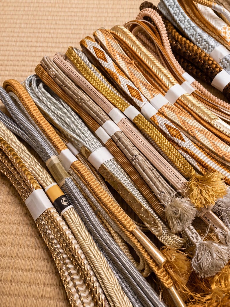 May include: A collection of intricately woven decorative cords in various shades of gold, silver, and beige. Some cords have tassels. The cords are bundled together with white paper bands, and are arranged on a light brown surface.
