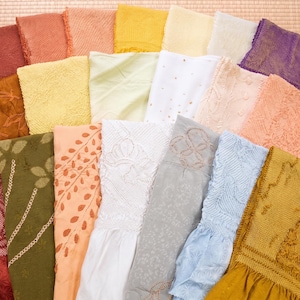 May include: A collection of colorful, folded fabric squares in various shades of red, orange, yellow, green, white, and blue. The fabrics feature diverse textures and patterns, including embroidery and floral designs, arranged on a light brown surface.