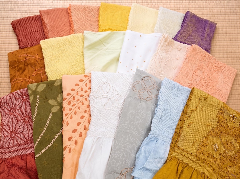 May include: A collection of colorful, folded fabric squares in various shades of red, orange, yellow, green, white, and blue. The fabrics feature diverse textures and patterns, including embroidery and floral designs, arranged on a light brown surface.