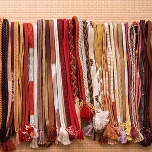 May include: A collection of colorful, decorative Japanese obi cords. The cords vary in color, texture, and design, including braided, patterned, and tasseled styles. Colors include red, brown, gold, and white. The cords are arranged vertically against a neutral background.